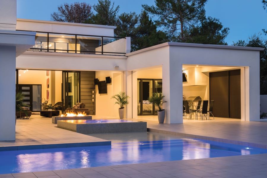 A backyard at dusk with a pool, fire feature, and an open-home concept with lights illuminating the home's interior. 