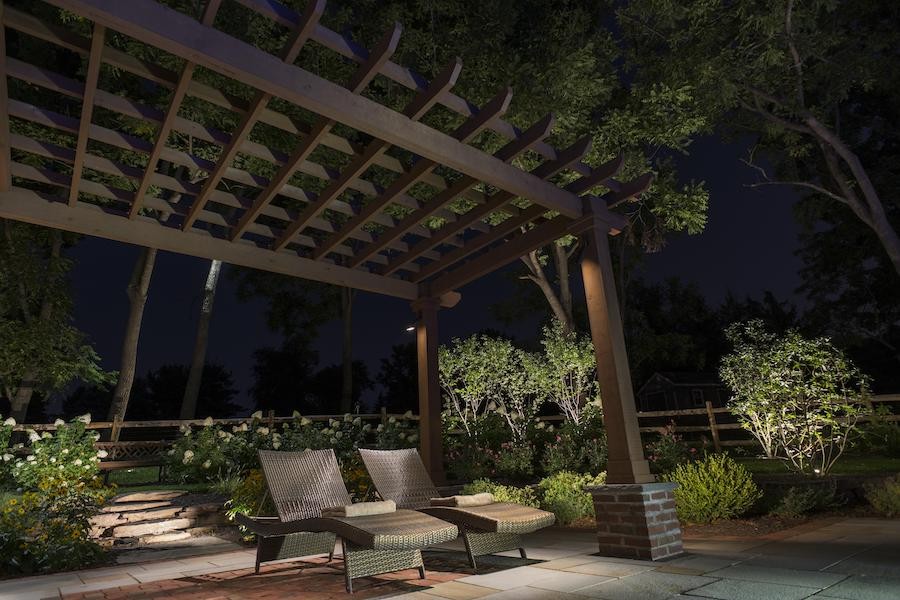 A backyard patio with seating is illuminated at nighttime with outdoor lighting.