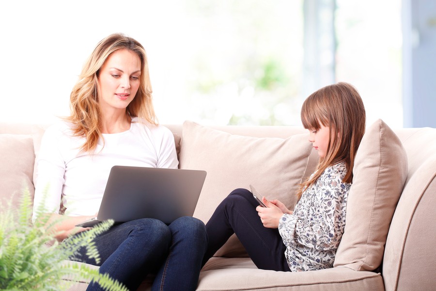 Woman and her daughter sitting on the couch, the woman with an open laptop on her lap and the child with a smart device. 