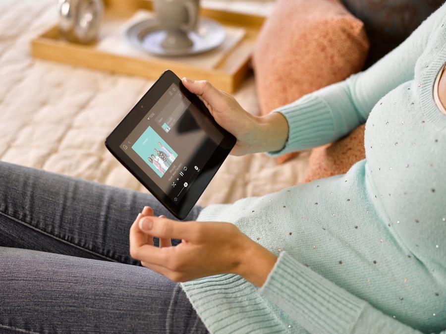 A woman sits on a couch and holds a tablet showing an album cover and music controls.