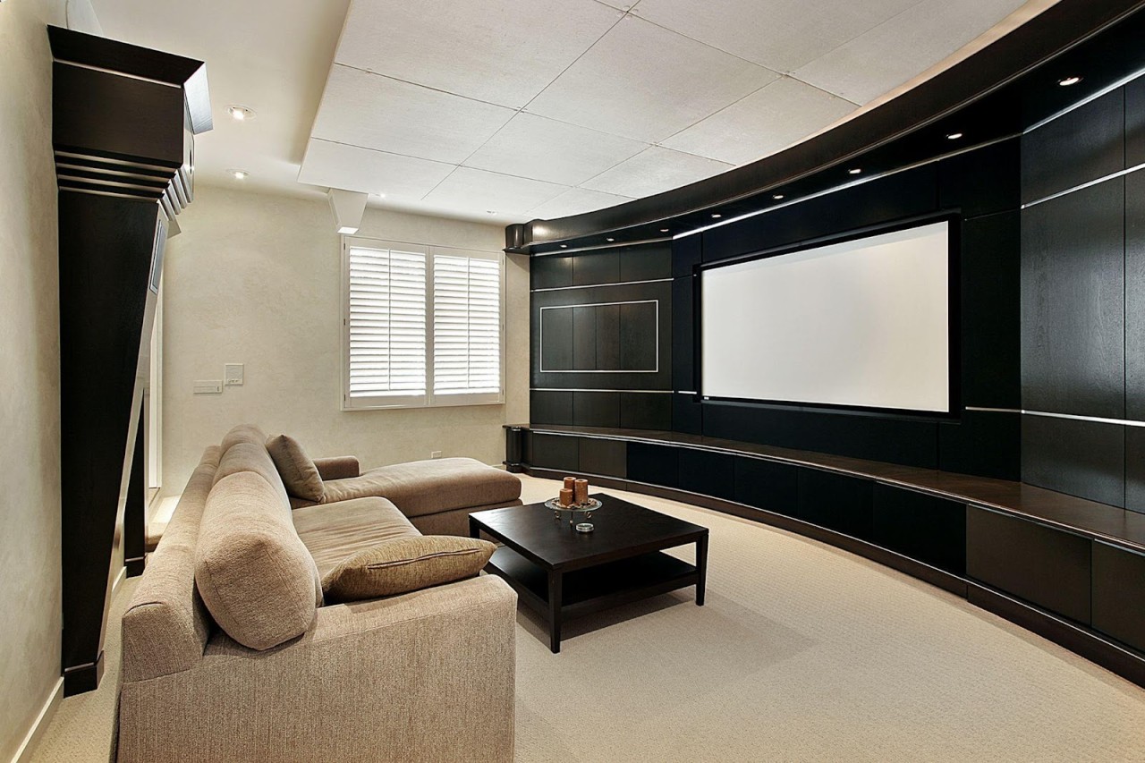 A beautiful home theater shown during the day with a beige couch and an enormous television.
